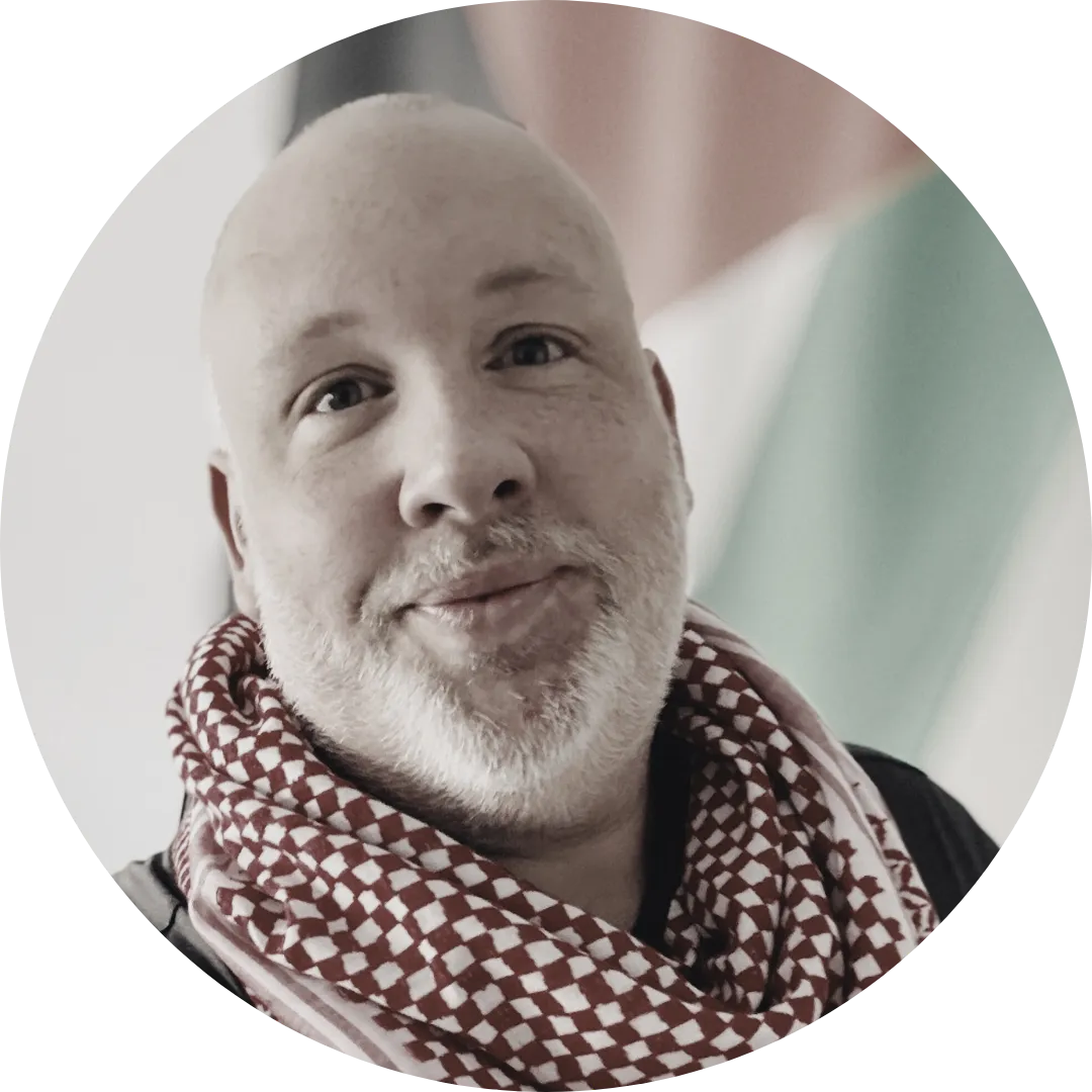 Portrait of Derron Borders, a chubby middle-aged person who was assigned male at birth who is bald with a greying beard. He is wearing a red kuffiyeh and the flag of Palestine hangs in the background.